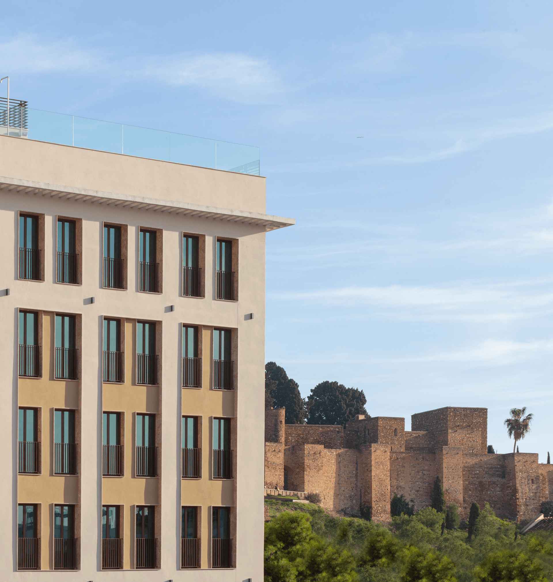 Lateral del edificio Hotel ME Málaga con la Alcazaba de Málaga al fondo bajo un cielo azul
